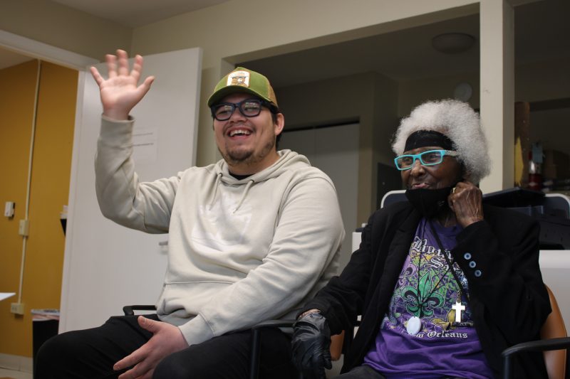 Man and older woman with glasses are sitting together. Man happily waves to the camera.
