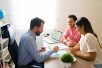 Couple seated at a desk with a man assisting them by reviewing documents in an office setting.
