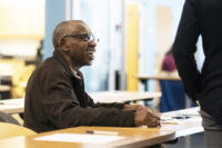 Older gentleman smiling and sitting in a workshop engaged with the teacher.