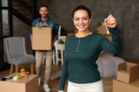 Woman smiling and holding a key while a man is in the background holding a cardboard box.